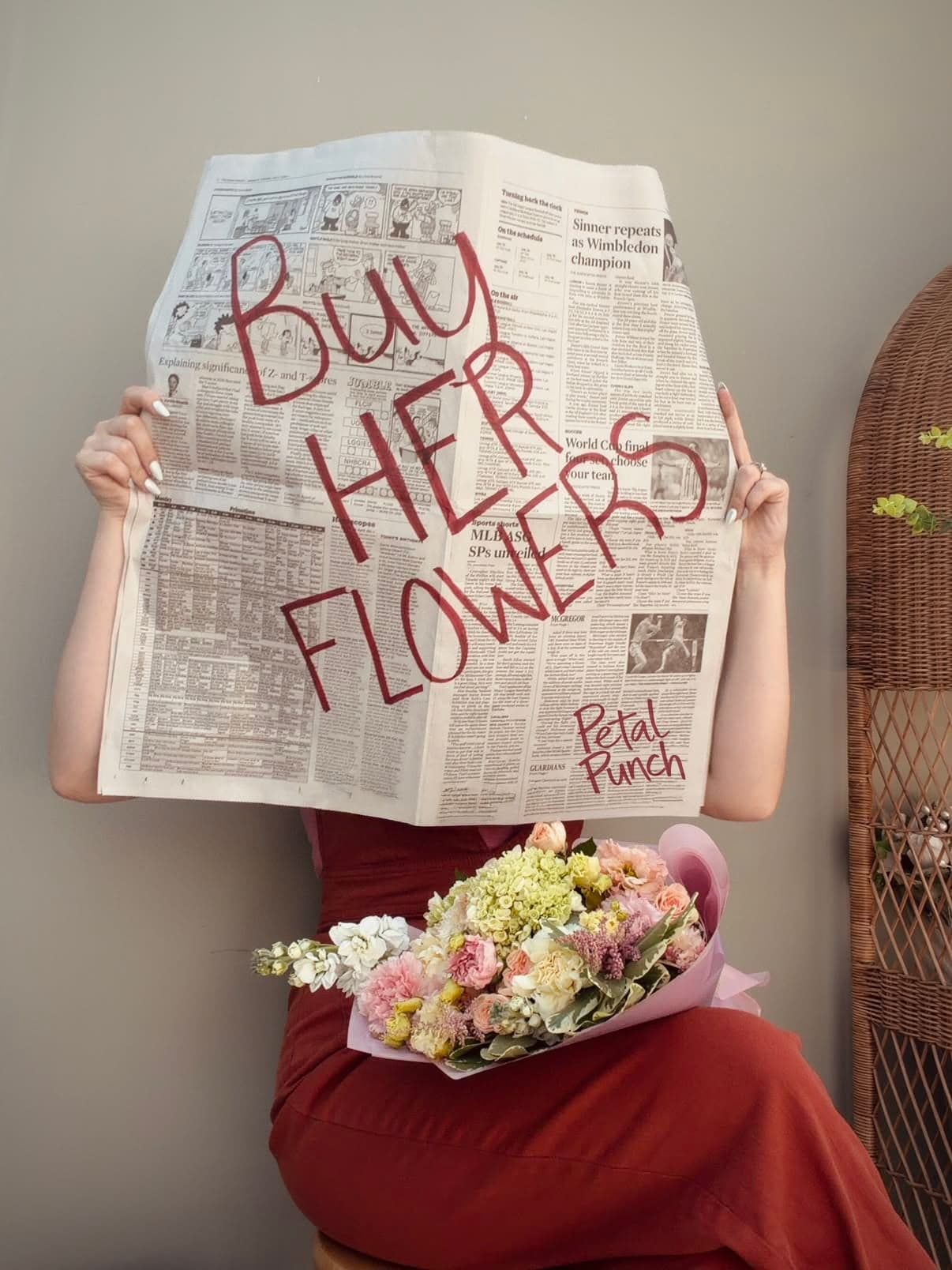 A woman in a rust dress hides behind a newspaper hand-lettered “Buy her flowers, Petal Punch”, a wrapped bouquet in her lap